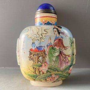 Puede incluir: Una botella de rapé con una escena pintada a mano de una figura junto a una mesa con incienso, rodeada de flores de loto. La botella tiene un cuerpo redondeado, una base de color claro y un tapón esférico azul con una banda metálica plateada.