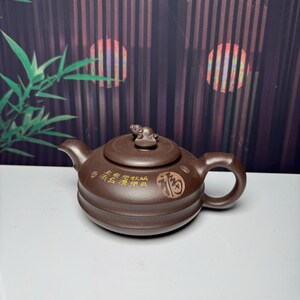 Könnte beinhalten: Eine Teekanne aus dunkelbraunem Ton mit rundem Körper, gebogenem Ausguss und kreisförmigem Griff. Auf dem Deckel befindet sich eine kleine, skulptierte Tierfigur. Die Seite der Kanne ist mit goldener chinesischer Kalligrafie und einem Siegel verziert.