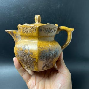 Puede incluir: Una tetera de cerámica color amarillo mostaza con textura moteada, cuerpo angular, tapa con un pequeño pomo y asa curva. El exterior está decorado con intrincados grabados oscuros de figuras y patrones ornamentales de volutas.