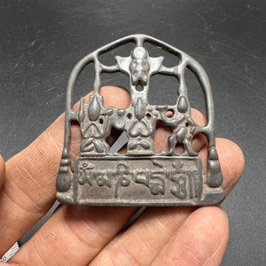 May include: A dark gray metal pendant featuring an arched frame with an openwork design. The center depicts three stylized figures, and the base contains a rectangular plaque with embossed Tibetan script characters in a relief style.