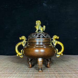 May include: A bronze-toned metal incense burner featuring a lid topped with a gold-colored lion figure. The vessel has two gold-colored dragon-shaped handles on the sides and stands on three legs shaped like elephant trunks with intricate details.