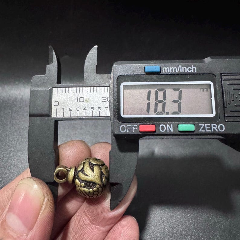 Puede incluir: Una peque&ntilde;a cuenta redonda de metal en color lat&oacute;n antiguo con un intrincado patr&oacute;n floral en relieve y un lazo superior. Un calibrador digital negro mide la cuenta, mostrando un ancho de 18,3 mil&iacute;metros en la pantalla digital clara.