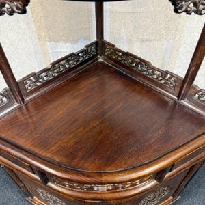 May include: A dark brown wooden corner cabinet featuring a triangular top surface, vertical support posts, and intricate openwork carvings along the edges. The piece includes a curved front panel with detailed relief carvings and a smooth finish.