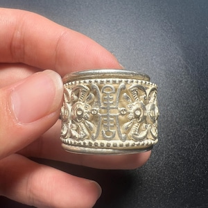 May include: A wide silver-toned ring featuring an intricate openwork design with repeating stylized figures and a central vertical symbol. The band is bordered by rows of small raised beads along the top and bottom edges, creating a textured finish.