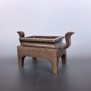 Pode incluir: Um queimador de incenso retangular de bronze com um acabamento met&aacute;lico liso e desgastado. O recipiente &eacute; suportado por quatro pernas robustas e inclui duas al&ccedil;as curvas que se estendem para cima, criando um design equilibrado e aberto.