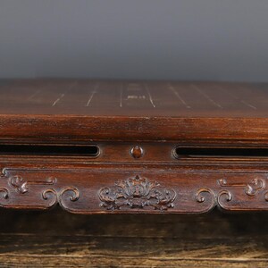May include: A dark brown wooden board with a smooth, flat top surface featuring thin, light-colored grid lines. The front edge of the board displays intricate, carved floral patterns and horizontal slots, showcasing a polished, deep-toned wood grain.