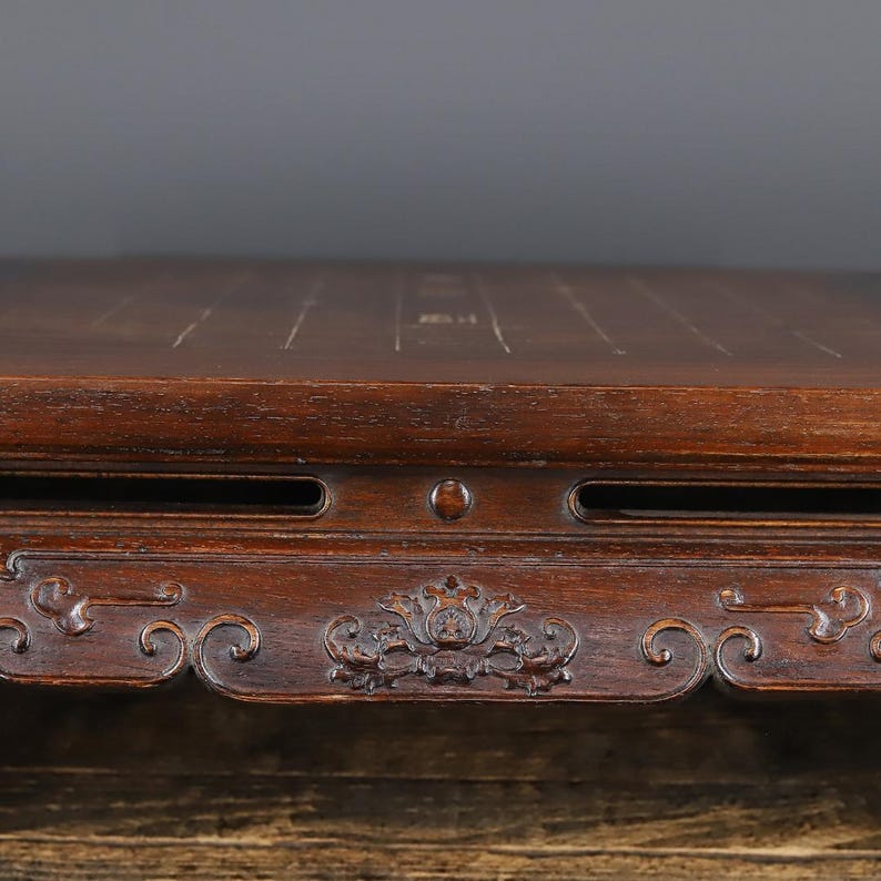 May include: A dark brown wooden board with a smooth, flat top surface featuring thin, light-colored grid lines. The front edge of the board displays intricate, carved floral patterns and horizontal slots, showcasing a polished, deep-toned wood grain.