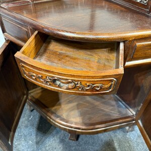 May include: A dark brown wooden corner cabinet featuring a curved front edge and a single pull-out drawer. The drawer front is decorated with carved scrollwork patterns and a central recessed handle. The cabinet has open side doors.