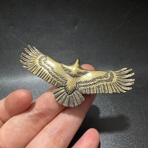 以下が含まれることがあります： 翼を大きく広げて飛ぶ鷲の形をした真鍮色の金属製ブローチ。翼と尾には羽の模様が細かく刻まれており、頭部と胴体の中央部分は滑らかで光沢のある仕上げになっています。