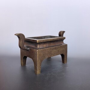 Pode incluir: Um queimador de incenso retangular de bronze com uma p&aacute;tina escura e desgastada, quatro pernas robustas e duas al&ccedil;as curvadas para cima nos lados opostos. O recipiente tem uma abertura larga e uma constru&ccedil;&atilde;o met&aacute;lica s&oacute;lida e uniforme.