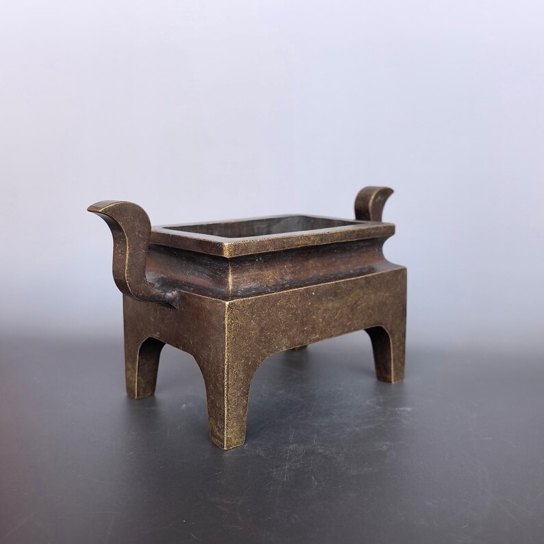Pode incluir: Um queimador de incenso retangular de bronze com uma p&aacute;tina escura e desgastada, quatro pernas robustas e duas al&ccedil;as curvadas para cima nos lados opostos. O recipiente tem uma abertura larga e uma constru&ccedil;&atilde;o met&aacute;lica s&oacute;lida e uniforme.