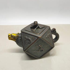 May include: A dark bronze-toned teapot with a cubic body featuring embossed animal figures on its sides. The spout is shaped like a dragon head with gold-colored accents, and the lid is topped with a small sculpted monkey figure.