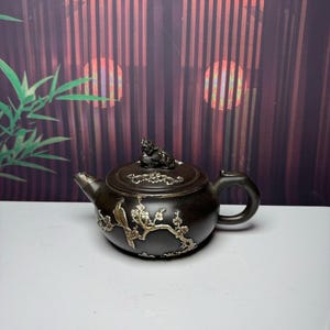 May include: A dark brown teapot featuring silver-toned metal inlays of a bird perched on a flowering branch. The lid is topped with a small sculpted animal figure, and the spout and handle are integrated into the rounded body of the vessel.