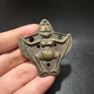 Pode incluir: Um pingente de metal em tom de bronze apresentando uma figura estilizada com um torso arredondado, apêndices em forma de asas estendidas e uma cabeça com três pequenas protuberâncias circulares no topo. A peça inclui quatro recortes circulares.