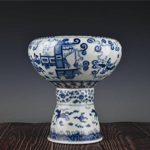 May include: A blue and white porcelain stem cup featuring intricate hand-painted scenes of figures in architectural settings, swirling cloud motifs, floral vines, and galloping horses around the flared base, finished with a glossy glaze.