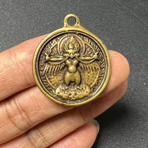 May include: A circular brass pendant featuring a raised relief of a multi-armed deity figure centred within a decorative border. The metal has a warm, aged gold tone with dark recessed areas that highlight the intricate details of the central figure.