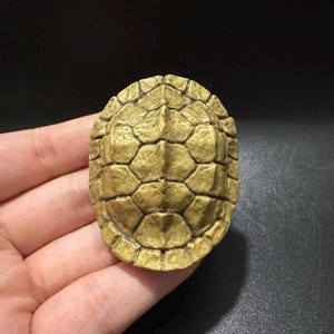Puede incluir: Una pequeña figura con forma de caparazón de tortuga ovalado con una superficie texturizada de color marrón dorado. El caparazón presenta un patrón distintivo de escudos hexagonales en relieve y una cresta central prominente.