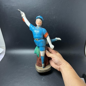 May include: A ceramic figurine of a person in a blue uniform and cap, holding a small black pistol in an outstretched arm. The figure wears a red armband with yellow characters, a brown belt, and stands on a brown base with green accents.