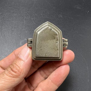 Puede incluir: Un pequeño relicario de metal plateado con una parte superior puntiaguda en forma de arco. El objeto presenta un panel central liso y elevado con un borde escalonado y dos bisagras laterales, formando una pieza compacta con acabado mate.