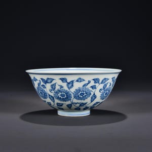 Op de afbeelding: Een witte keramische kom met een handgeschilderd blauw bloemmotief van gedetailleerde chrysantenbloesems en krullende wijnbladeren. De kom heeft een uitlopende rand en een ronde voet, met dunne blauwe lijnen rondom de onderkant.
