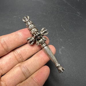 May include: A silver-toned metal phurba ritual dagger featuring a handle adorned with three stacked deity faces, a central cross-guard with claw-like protrusions, and a tapered, smooth blade section decorated with etched spiral patterns at the base.