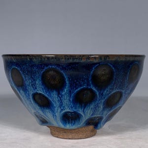 May include: A ceramic bowl featuring a deep blue glaze with dark, circular, oil-spot patterns that bleed into lighter blue streaks. The bowl has a smooth, curved rim and a thick, unglazed, light brown clay foot at the base, providing a contrast.