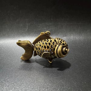 May include: A brass-toned metal fish pendant featuring an intricate openwork lattice pattern across the body, a solid textured tail, and a small circular loop at the top for attachment. The design includes detailed fins and a prominent eye.