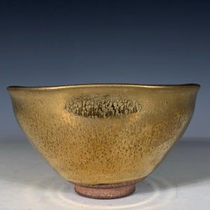 Könnte beinhalten: Eine Keramikschale mit metallisch goldener Glasur, die eine gesprenkelte Textur auf der Außenfläche aufweist. Die Schale hat einen glatten, geschwungenen Rand und steht auf einem markanten, unglasierten, rotbraunen runden Fuß mit rauer Oberfläche.