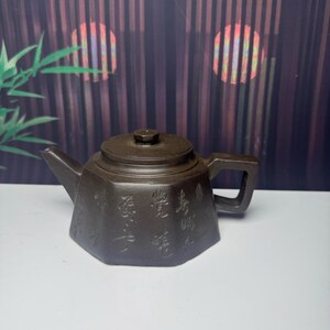May include: A dark brown hexagonal teapot with a flat lid and a square-shaped handle. The body of the teapot features engraved Chinese calligraphy characters on its front side. The spout is short and angled, extending from the main vessel.