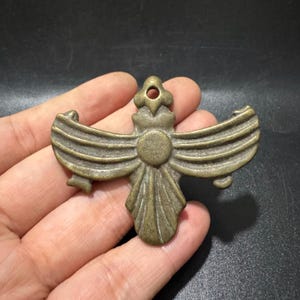 Può includere: Un ciondolo in metallo color bronzo raffigurante un emblema alato stilizzato con un disco circolare centrale, ali curve simmetriche con creste orizzontali, una base a ventaglio e un piccolo occhiello circolare nella parte superiore.