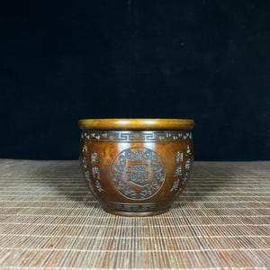 Pode incluir: Uma taça redonda de bronze castanho escuro com um rebordo liso e polido. O exterior apresenta um medalhão circular central com caracteres chineses em relevo, rodeado por volutas intrincadas e colunas verticais de texto chinês gravado.