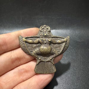 Può includere: Un ciondolo in metallo color bronzo raffigurante una figura alata stilizzata con busto arrotondato, ali spiegate e coda a ventaglio. La figura presenta un volto dettagliato con un copricapo e piume testurizzate sulle ali.