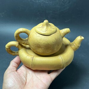 Puede incluir: Una tetera de cerámica de tono amarillo con una superficie texturizada de patrón agrietado y una tapa a juego, que descansa sobre una base circular de cerámica con forma de bambú. La tetera presenta un asa curva y un pico vertedor.