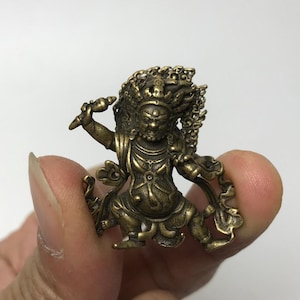 Pode incluir: Pequena estatueta de cor bronze de uma divindade em pose dinâmica. A figura tem um cocar detalhado e segura uma arma. O design geral sugere um tema religioso ou espiritual, com detalhes intrincados e uma aparência desgastada.