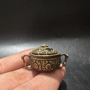 May include: A small, round, antique-style brass incense burner featuring two ornate side handles and a removable lid. The exterior is decorated with intricate embossed patterns, including circular motifs and a geometric border along the top rim.