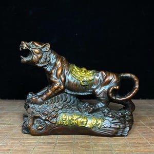 Könnte beinhalten: Eine bronzefarbene Tigerfigur in lauernder Haltung mit geöffnetem Maul und einem goldfarbenen Sattel auf dem Rücken. Der Sockel ist wie Felsen und Münzen geformt und trägt auf der Vorderseite goldfarbene chinesische Schriftzeichen.