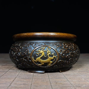 Pode incluir: Um queimador de incenso de metal em tom de bronze escuro com um medalhão circular apresentando um motivo de dragão em relevo na cor dourada. A superfície externa é decorada com padrões de nuvens e o recipiente possui uma borda lisa.