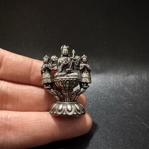 May include: A small silver-toned metal figurine depicting a central seated figure holding a sword, flanked by two smaller standing figures. The entire piece is mounted on a decorative lotus-shaped base with intricate petal detailing throughout.