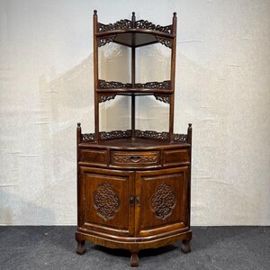 May include: A dark brown wooden corner cabinet featuring an open upper section with two shelves and intricate carved detailing. The base includes a central drawer and two lower cabinet doors, each adorned with a symmetrical carved floral motif.