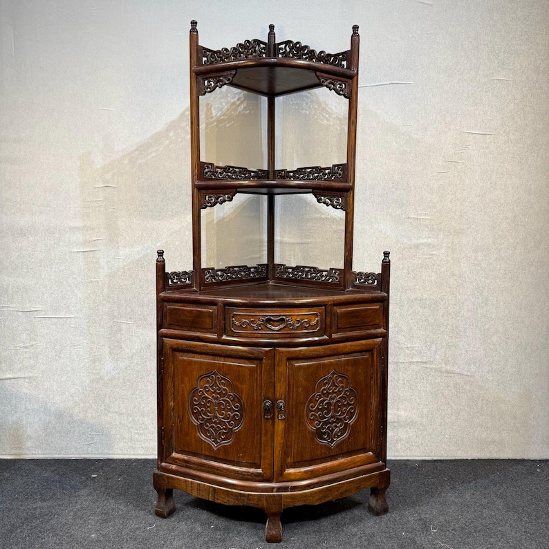 May include: A dark brown wooden corner cabinet featuring an open upper section with two shelves and intricate carved detailing. The base includes a central drawer and two lower cabinet doors, each adorned with a symmetrical carved floral motif.