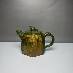 Puede incluir: Una tetera de cerámica verde oscuro con un diseño en relieve de color dorado que muestra una grulla entre árboles. La tetera tiene un asa curva, un pico con forma de cabeza de animal y una tapa rematada con un pequeño remate de ave.