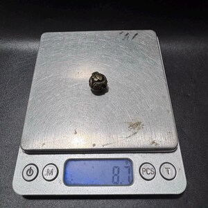 Puede incluir: Una peque&ntilde;a cuenta esf&eacute;rica de metal oscuro con un intrincado patr&oacute;n floral o de volutas en relieve. La cuenta est&aacute; centrada en una b&aacute;scula digital que muestra un peso de 8,7 gramos, proporcionando una referencia visual de su masa.