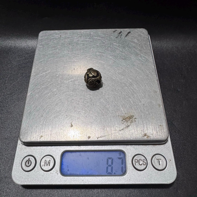 Puede incluir: Una peque&ntilde;a cuenta esf&eacute;rica de metal oscuro con un intrincado patr&oacute;n floral o de volutas en relieve. La cuenta est&aacute; centrada en una b&aacute;scula digital que muestra un peso de 8,7 gramos, proporcionando una referencia visual de su masa.