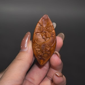 May include: A brown, almond-shaped wooden pendant featuring an intricate, relief-carved dragon design. The carving displays detailed scales, a serpentine body, and a horned head, all contained within the natural, pointed contours of the wood piece.