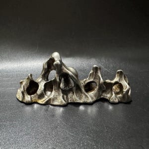 May include: A silver-toned metal object shaped like a jagged mountain range with multiple hollow openings and a textured, uneven surface. The piece features several peaks and valleys, creating a porous, organic form with a metallic, weathered finish.