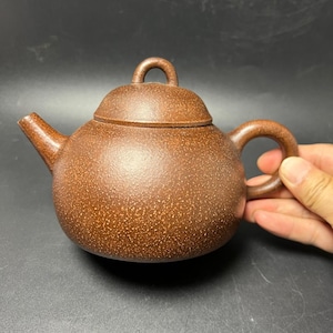 Puede incluir: Una tetera redonda de color marrón con una superficie texturizada y moteada. La tetera presenta un pico curvo, un asa circular y una tapa a juego con un pequeño mango arqueado en la parte superior. El cuerpo es esférico y tiene un acabado mate.