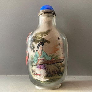 Puede incluir: Una botella de rapé de vidrio transparente con una pintura interior de una persona con ropa tradicional tocando un instrumento de cuerda bajo una palmera. La botella tiene un tapón azul redondeado con un borde de metal plateado y caracteres chinos negros.