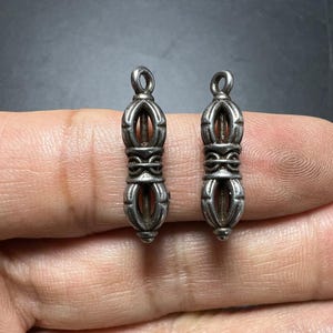 May include: Two small, silver-toned metal pendants shaped like vajras. Each piece features a central cylindrical section with symmetrical, curved prongs extending from the top and bottom, and a small circular loop at the very top for hanging.