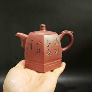 Puede incluir: Una tetera de arcilla de color marrón rojizo con forma cuadrada, que presenta una tapa con un pequeño pomo, un asa curva y un pico corto. La superficie exterior está decorada con caracteres de caligrafía china grabados en oscuro.