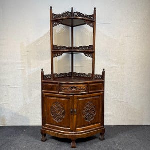 May include: A dark brown wooden corner cabinet featuring an open upper section with two triangular shelves and intricate carved lattice detailing. The base includes a central drawer and two lower cabinet doors with ornate carved circular motifs.